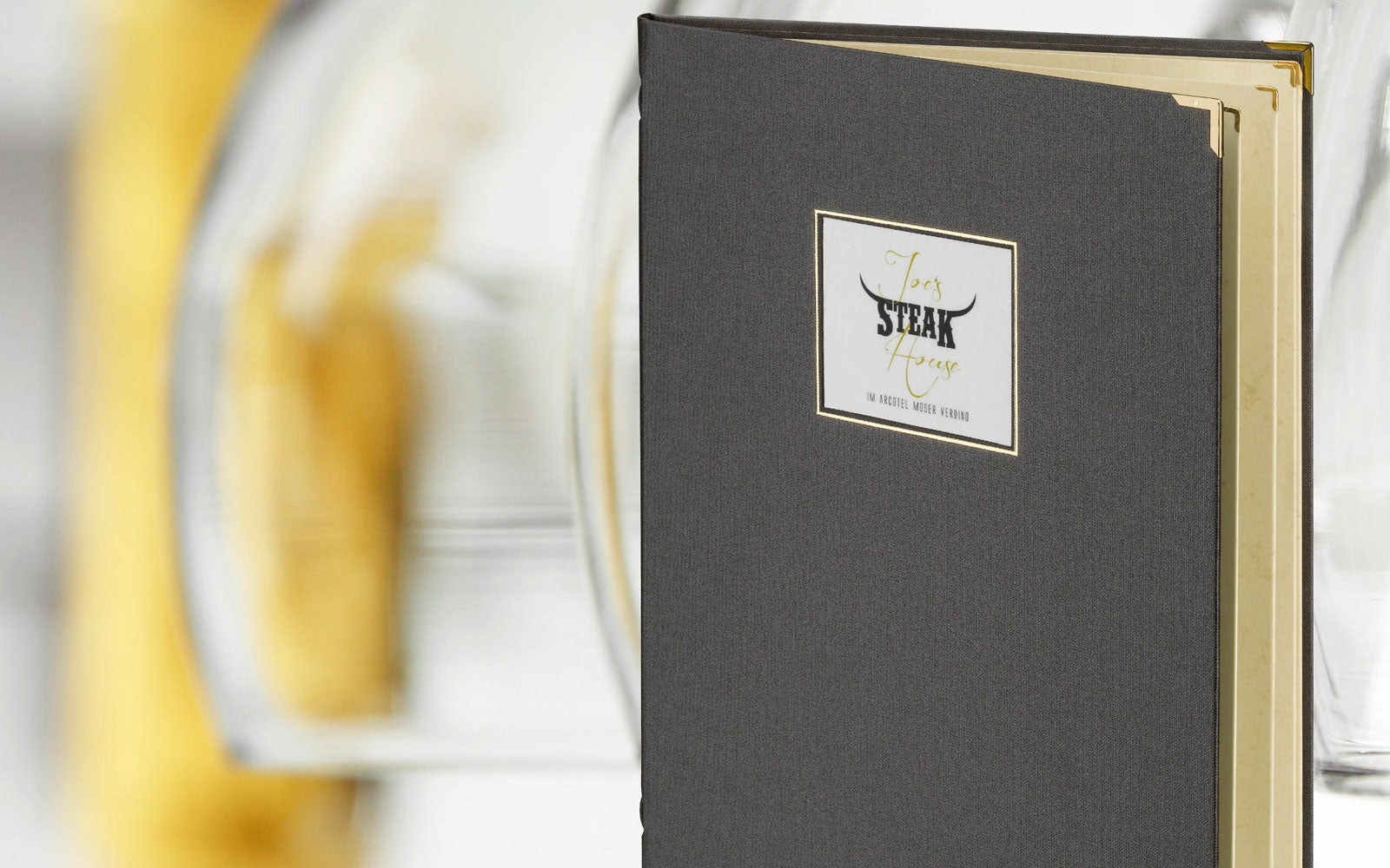 Detailaufnahme einer Speisekarte GrandXXL aus Buchleinen in der Farbe grau mit Veredelung auf der Vorderseite und Metallecken in Gold